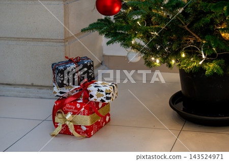 Wrapped Christmas Gifts on Floor by Potted Tree Base. Festive wrapped gifts including a red Merry Christmas box stacked on the floor beside the base of a potted Christmas tree with fairy lights. Wrapped Christmas Gifts on Floor by Potted Tree Base. Festive wrapped gifts including a red Merry Christmas box stacked on the floor beside the base of a potted Christmas tree with fairy lights. 143524971