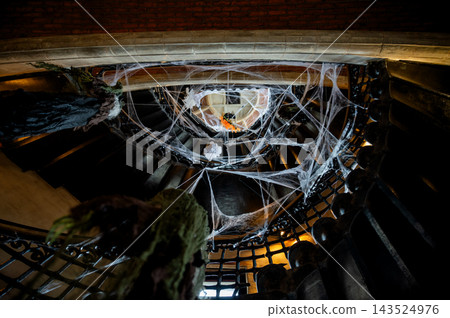 Cobweb Draped Staircase with Spooky Halloween Decor. White cobwebs drape a dark vintage staircase with hanging decorations creating a haunted Halloween atmosphere. An eerie indoor scene of festive Cobweb Draped Staircase with Spooky Halloween Decor. White cobwebs drape a dark vintage staircase with hanging decorations creating a haunted Halloween atmosphere. An eerie indoor scene of festive 143524976