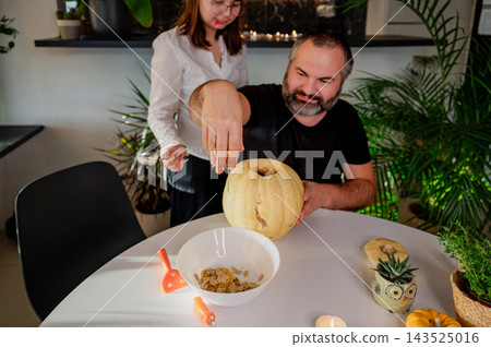 Father Emptying Hollowed Pumpkin over Bowl for Halloween. Her father holds a hollowed pale pumpkin over a bowl while a daughter stands behind him at a bright festive table with mini pumpkins and a wit Father Emptying Hollowed Pumpkin over Bowl for Halloween. Her father holds a hollowed pale pumpkin over a bowl while a daughter stands behind him at a bright festive table with mini pumpkins and a wit 143525016