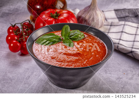 Gray stone surface with black bowl holding red gazpacho topped with fresh basil leaves beside cherry tomatoes and garlic bulb as cold soup dish Gray stone surface with black bowl holding red gazpacho topped with fresh basil leaves beside cherry tomatoes and garlic bulb as cold soup dish 143525234