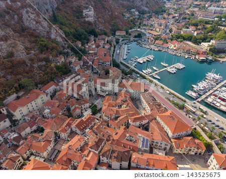 Kotor Montenegro coastal town spreads around sheltered marina, seen from above. Red tiled roofs and pale stone houses line harbor roads, while sailboats sit among piers beneath rugged slopes. Kotor Montenegro coastal town spreads around sheltered marina, seen from above. Red tiled roofs and pale stone houses line harbor roads, while sailboats sit among piers beneath rugged slopes. 143525675