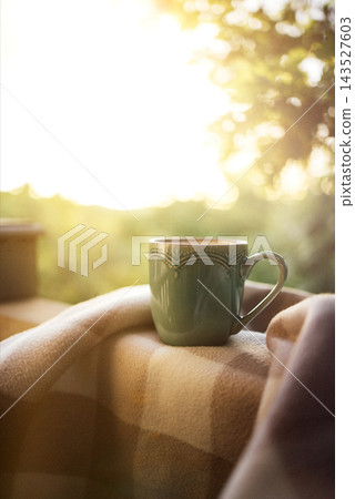 Green ceramic mug resting on a warm plaid blanket beside a sunlit window, with soft golden outdoor foliage creating a tranquil cozy still life Green ceramic mug resting on a warm plaid blanket beside a sunlit window, with soft golden outdoor foliage creating a tranquil cozy still life 143527603