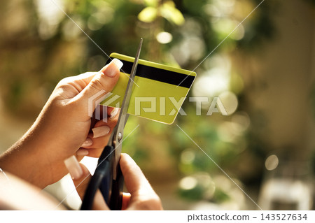 Hands cutting a blank yellow and black plastic card with scissors against softly blurred green foliage and warm sunlight, closeup household object scene 143527634