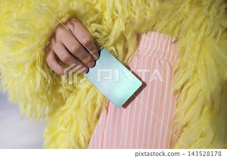 Female hand holding a reflective card while wearing a bright yellow fluffy coat and pink striped pants, showcasing a vibrant fashion style in a contemporary setting Female hand holding a reflective card while wearing a bright yellow fluffy coat and pink striped pants, showcasing a vibrant fashion style in a contemporary setting 143528178
