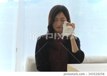 一位年輕女子站在窗邊,用紙巾擦眼淚,表達悲傷(悲傷、眼淚、震驚和痛苦的畫面)。 一位年輕女子站在窗邊,用紙巾擦眼淚,表達悲傷(悲傷、眼淚、震驚和痛苦的畫面)。 143528256