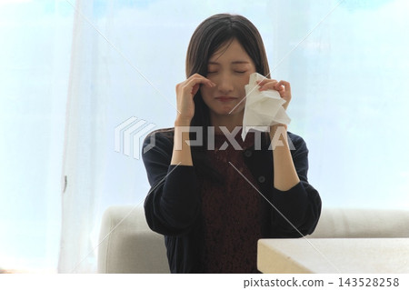 一位年輕女子站在窗邊,用紙巾擦眼淚,表達悲傷(悲傷、眼淚、震驚和痛苦的畫面)。 一位年輕女子站在窗邊,用紙巾擦眼淚,表達悲傷(悲傷、眼淚、震驚和痛苦的畫面)。 143528258