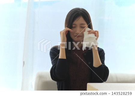 一位年輕女子站在窗邊,用紙巾擦眼淚,表達悲傷(悲傷、眼淚、震驚和痛苦的畫面)。 一位年輕女子站在窗邊,用紙巾擦眼淚,表達悲傷(悲傷、眼淚、震驚和痛苦的畫面)。 143528259