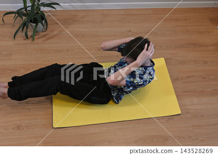 Boy does an exercise on a yellow mat in a room with a green plant nearby during the day Boy does an exercise on a yellow mat in a room with a green plant nearby during the day 143528269