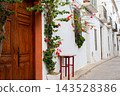 Quaint Altea street view in Spain, highlighting traditional white architecture, colorful bougainvillea, a rustic wooden door, and a small outdoor table, perfect for family holidays and tourism 143528386