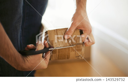 Hands cutting open sealed cardboard parcel with scissors beside bright window, close view of delivery package inspection at home on wooden surface Hands cutting open sealed cardboard parcel with scissors beside bright window, close view of delivery package inspection at home on wooden surface 143529732