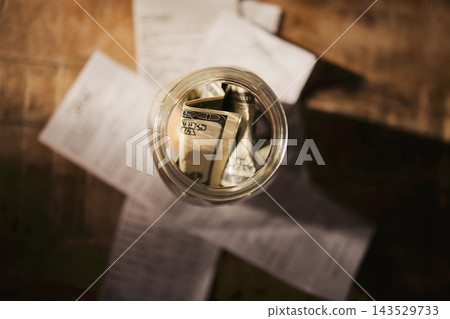 Top view of glass jar containing folded banknotes over scattered printed papers on rustic wooden table, warm natural light and muted earthy tones, still life photograph 143529733
