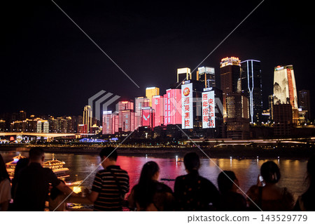 City skyline at night featuring illuminated buildings and colorful advertisements along the riverbank with people enjoying the view in Chongqing, China 143529779