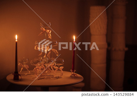 Elegant glass champagne tower with pink ribbons and two lit candles on a round table, soft ambient lighting creates a romantic atmosphere in a cozy setting Elegant glass champagne tower with pink ribbons and two lit candles on a round table, soft ambient lighting creates a romantic atmosphere in a cozy setting 143529884