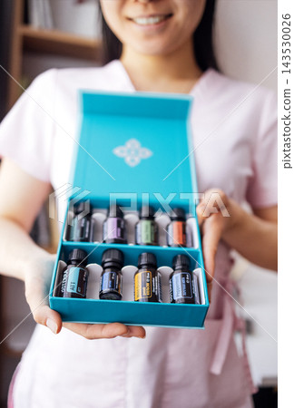 Asian woman in pink attire holds open a blue box containing various essential oil bottles, showcasing a collection of aromatic products for wellness and relaxation Asian woman in pink attire holds open a blue box containing various essential oil bottles, showcasing a collection of aromatic products for wellness and relaxation 143530026