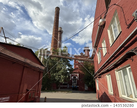 Historic red brick industrial courtyard with tall smokestacks, overhead pipes, blue sky, and clouds Historic red brick industrial courtyard with tall smokestacks, overhead pipes, blue sky, and clouds 143530280