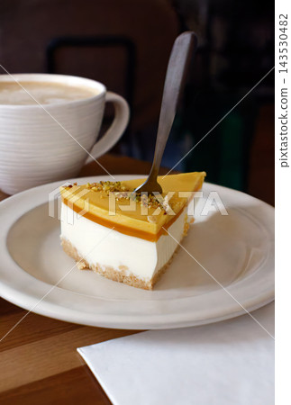 Slice of delicious cheesecake topped with yellow jelly and chopped pistachios served on a white plate with a coffee cup in the background on a wooden table in a cafe. Slice of delicious cheesecake topped with yellow jelly and chopped pistachios served on a white plate with a coffee cup in the background on a wooden table in a cafe. 143530482