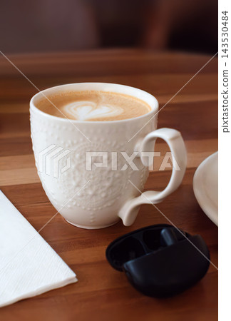 White ceramic cup of cappuccino with heart shaped latte art foam served on a wooden table next to wireless earphones case and napkin in a cafe. Concept of work break and relaxation. White ceramic cup of cappuccino with heart shaped latte art foam served on a wooden table next to wireless earphones case and napkin in a cafe. Concept of work break and relaxation. 143530484