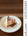 Slice of delicious cheesecake topped with caramel sauce and coconut flakes on a white plate with a fork. Sweet dessert served on wooden table in cafe. 143530485