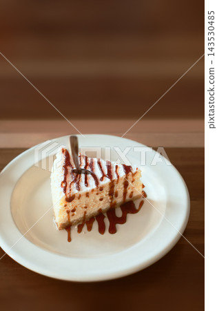 Slice of delicious cheesecake topped with caramel sauce and coconut flakes on a white plate with a fork. Sweet dessert served on wooden table in cafe. Slice of delicious cheesecake topped with caramel sauce and coconut flakes on a white plate with a fork. Sweet dessert served on wooden table in cafe. 143530485