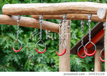 Children play on red hanging rings attached to a wooden outdoor playground. Play, fitness, childhood, park activity, obstacle course, coordination, recreation. Children play on red hanging rings attached to a wooden outdoor playground. Play, fitness, childhood, park activity, obstacle course, coordination, recreation. 143531129