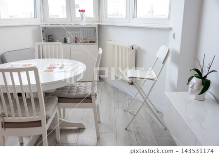 Bright minimalist dining and living area with white furniture and a potted plant by the window. Interior design, minimalism, home lifestyle, calm, simplicity, cozy morning. Bright minimalist dining and living area with white furniture and a potted plant by the window. Interior design, minimalism, home lifestyle, calm, simplicity, cozy morning. 143531174