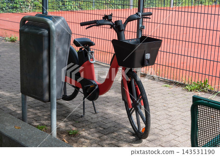 Red shared electric bike parked beside a bench near a playground in a city park. Urban mobility, shared transport, sustainability, commuting, family leisure, outdoor recreation. Red shared electric bike parked beside a bench near a playground in a city park. Urban mobility, shared transport, sustainability, commuting, family leisure, outdoor recreation. 143531190