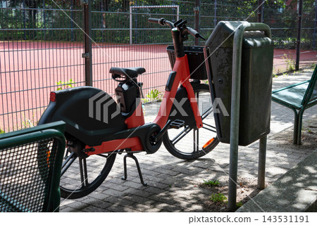 Red shared electric bike parked beside a bench near a playground in a city park. Urban mobility, shared transport, sustainability, commuting, family leisure, outdoor recreation. Red shared electric bike parked beside a bench near a playground in a city park. Urban mobility, shared transport, sustainability, commuting, family leisure, outdoor recreation. 143531191