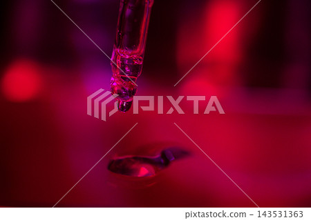 Transparent glass dropper releasing liquid droplet above reflective surface, illuminated by vivid red and purple light with blurred background and dramatic close up still life Transparent glass dropper releasing liquid droplet above reflective surface, illuminated by vivid red and purple light with blurred background and dramatic close up still life 143531363