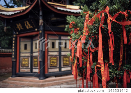 Red ribbons tied to a tree near a traditional Chinese temple with ornate architecture and gold inscriptions visible on the building's exterior 143531442