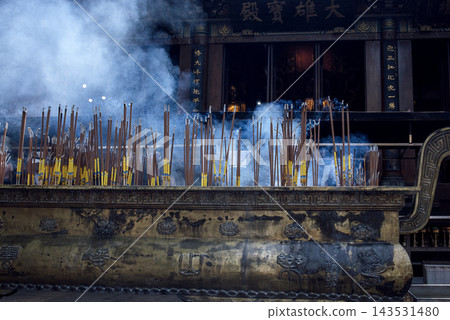 Incense sticks burning in a large bronze holder, smoke rising in front of a temple with ornate decorations and statues visible in the background Incense sticks burning in a large bronze holder, smoke rising in front of a temple with ornate decorations and statues visible in the background 143531480