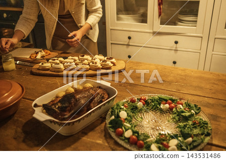Woman preparing festive meal with roasted meat, decorated platter, and assorted cookies on wooden table in cozy kitchen setting Woman preparing festive meal with roasted meat, decorated platter, and assorted cookies on wooden table in cozy kitchen setting 143531486