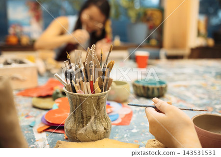 Art studio scene featuring a close-up of a ceramic pot filled with paintbrushes, with a blurred artist working on pottery in the background Art studio scene featuring a close-up of a ceramic pot filled with paintbrushes, with a blurred artist working on pottery in the background 143531531