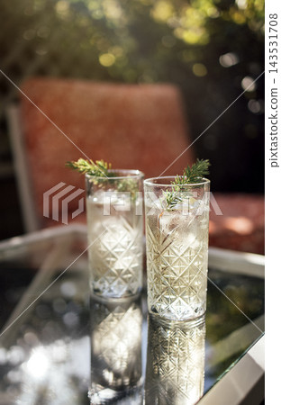 Two tall glasses of refreshing cocktail with ice and garnished with herbs placed on a glass table in a sunny outdoor setting with blurred background Two tall glasses of refreshing cocktail with ice and garnished with herbs placed on a glass table in a sunny outdoor setting with blurred background 143531708