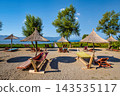 Sunny beach resort with lounge chairs and umbrellas by a lake with mountains in the background. 143535117