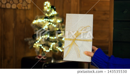 Person holds wrapped holiday gift box in front of illuminated Christmas tree, offering cheerful seasonal present decorated with golden ribbon and subtle glitter during festive celebration. 143535530