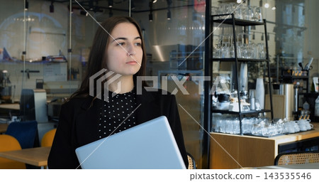 Young businesswoman holds closed modern laptop while standing inside bright modern coffee shop. Focused professional worker pauses with thoughtful expression during busy workday morning. Young businesswoman holds closed modern laptop while standing inside bright modern coffee shop. Focused professional worker pauses with thoughtful expression during busy workday morning. 143535546