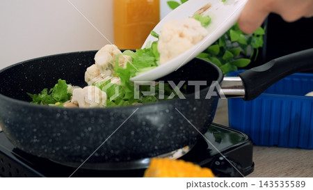 Person slides fresh cauliflower into frying pan during vegetarian meal prep routine indoors, adding leafy greens for nourishing stir fry dish while focused on balanced healthy nutrition daily. Person slides fresh cauliflower into frying pan during vegetarian meal prep routine indoors, adding leafy greens for nourishing stir fry dish while focused on balanced healthy nutrition daily. 143535589