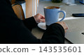 Person takes peaceful remote work break holding hot coffee mug on saucer near laptop computer. Calm worker rests hands beside ceramic cup on bright kitchen counter during morning pause. 143535601