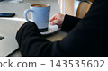 Young person adjusts ceramic mug filled with warm cappuccino while seated at desk. Female professional enjoys cozy morning coffee routine beside modern laptop during peaceful workday start. 143535602