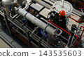Worker adjusts industrial pipe valve in busy plant, wearing red helmet and gloves. Maintenance worker stands on metal platform amid stainless steel piping and large equipment in industrial setting. 143535603