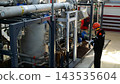 Focused young adult technician records notes while inspecting industrial machinery in futuristic facility. Technician stands near pipes, gauges, and control boxes under bright factory lighting. 143535604
