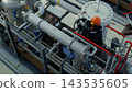Industrial worker tightens valve on pipe rack in industrial plant, focused and cautious in mechanical style. Worker stands amid stainless piping, gauges, and safety railings while using hand tools 143535605