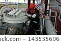 Crouched worker turns valve at refinery equipment, wearing red hard hat and goggles in industrial style. Worker grips tool beside bolted tank, pipelines, and metal railings with careful focus. 143535606