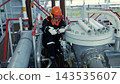Factory worker services heavy equipment with wrench, wearing hard hat, goggles, and gloves, in serious pose. Maintenance technician kneels among metal pipes, valves, and industrial machinery indoors. 143535607