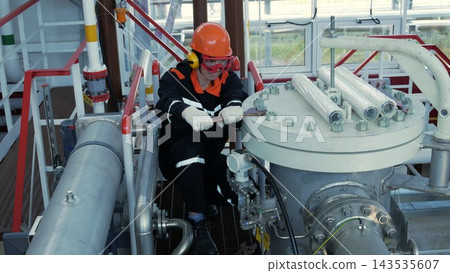 Factory worker services heavy equipment with wrench, wearing hard hat, goggles, and gloves, in serious pose. Maintenance technician kneels among metal pipes, valves, and industrial machinery indoors. 143535607