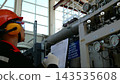 Protective gear worker fills inspection form near heavy industrial equipment, using clipboard and pen with focused expression. Maintenance worker faces pressure gauges and piping in spacious factory 143535608