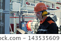 Focused male technician records notes on clipboard inside industrial facility, wearing hard hat, safety glasses, and earmuffs. Worker stands beside pipes, tanks, and ducts during plant inspection. 143535609