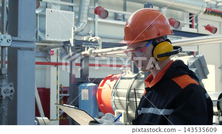 Focused male technician records notes on clipboard inside industrial facility, wearing hard hat, safety glasses, and earmuffs. Worker stands beside pipes, tanks, and ducts during plant inspection. Focused male technician records notes on clipboard inside industrial facility, wearing hard hat, safety glasses, and earmuffs. Worker stands beside pipes, tanks, and ducts during plant inspection. 143535609