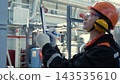 Focused technician in hard hat records inspection notes on clipboard inside industrial facility. Technician wearing gloves, goggles, and hearing protection stands near pipes and machinery. 143535610