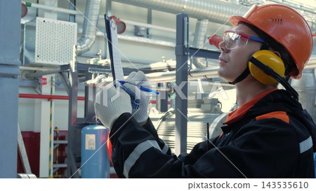 Focused technician in hard hat records inspection notes on clipboard inside industrial facility. Technician wearing gloves, goggles, and hearing protection stands near pipes and machinery. Focused technician in hard hat records inspection notes on clipboard inside industrial facility. Technician wearing gloves, goggles, and hearing protection stands near pipes and machinery. 143535610